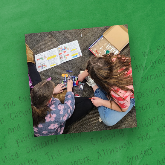 Two students sitting on the floor building electrical circuits from a kit, green textured background surrounds the photo